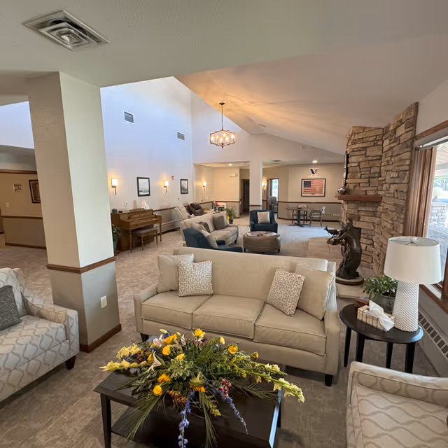 Bright, airy senior living lounge with beige sofas, stone fireplace, and floral centerpiece.