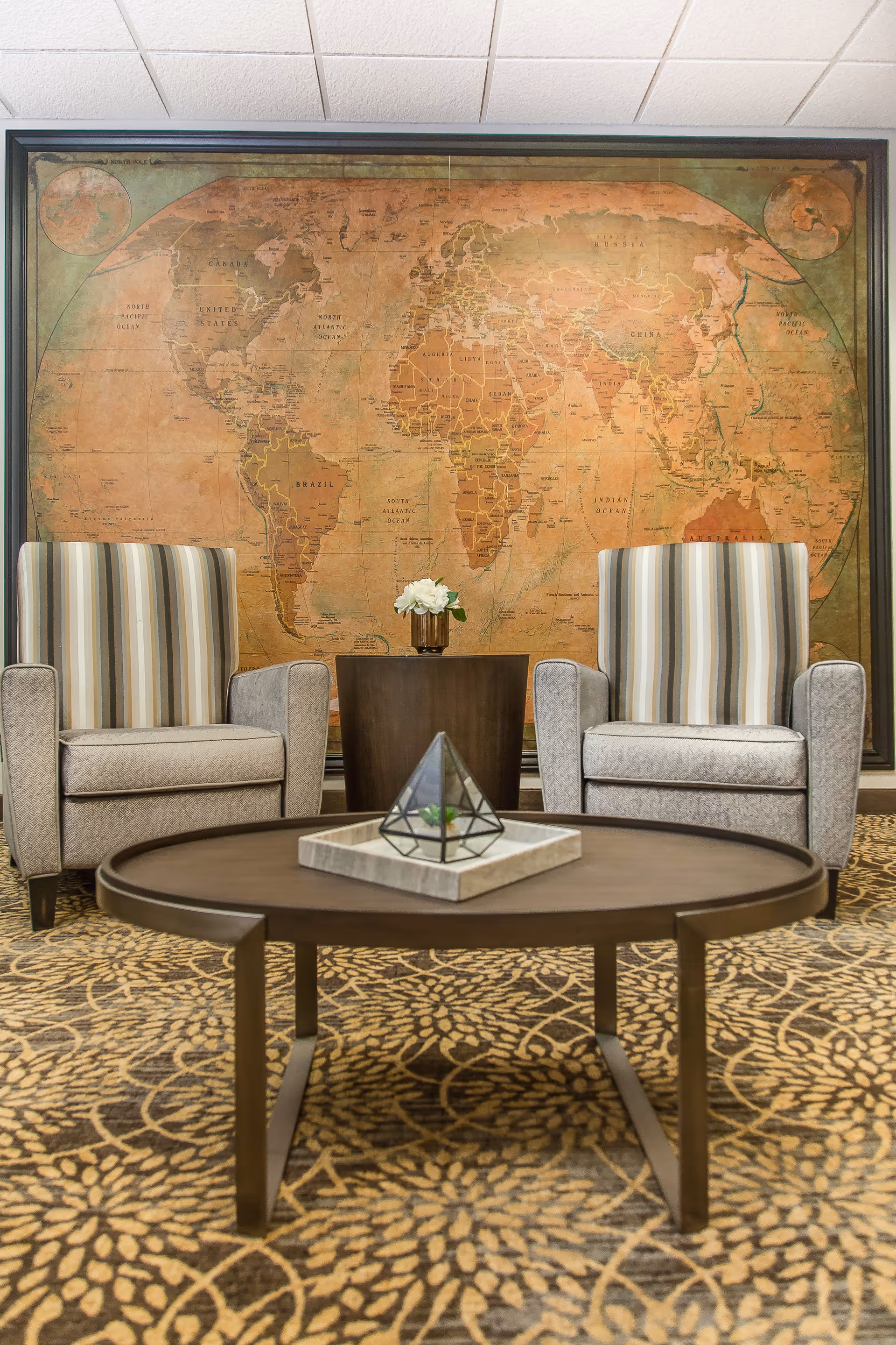 A cozy seating area with two striped armchairs on either side of a small round wooden table holding a vase with white flowers. In front of the chairs is a round coffee table with a decorative geometric terrarium on a tray. The background features a large vintage-style world map framed on the wall. The floor is covered with a patterned carpet.