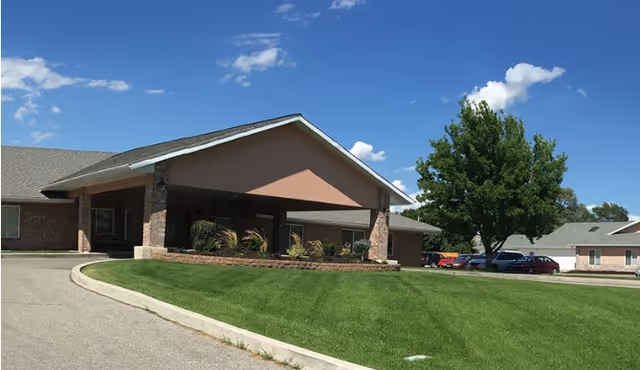 Front exterior of a single-story senior living building with a covered entrance, green lawn, and a tree under a blue sky.