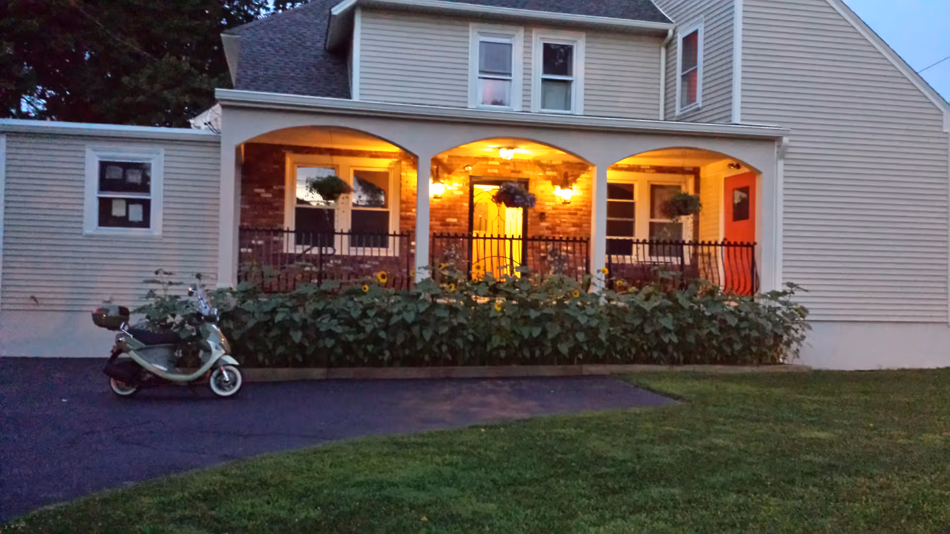 Two-story beige building with lit front porch, hedges, and a parked scooter in the driveway.