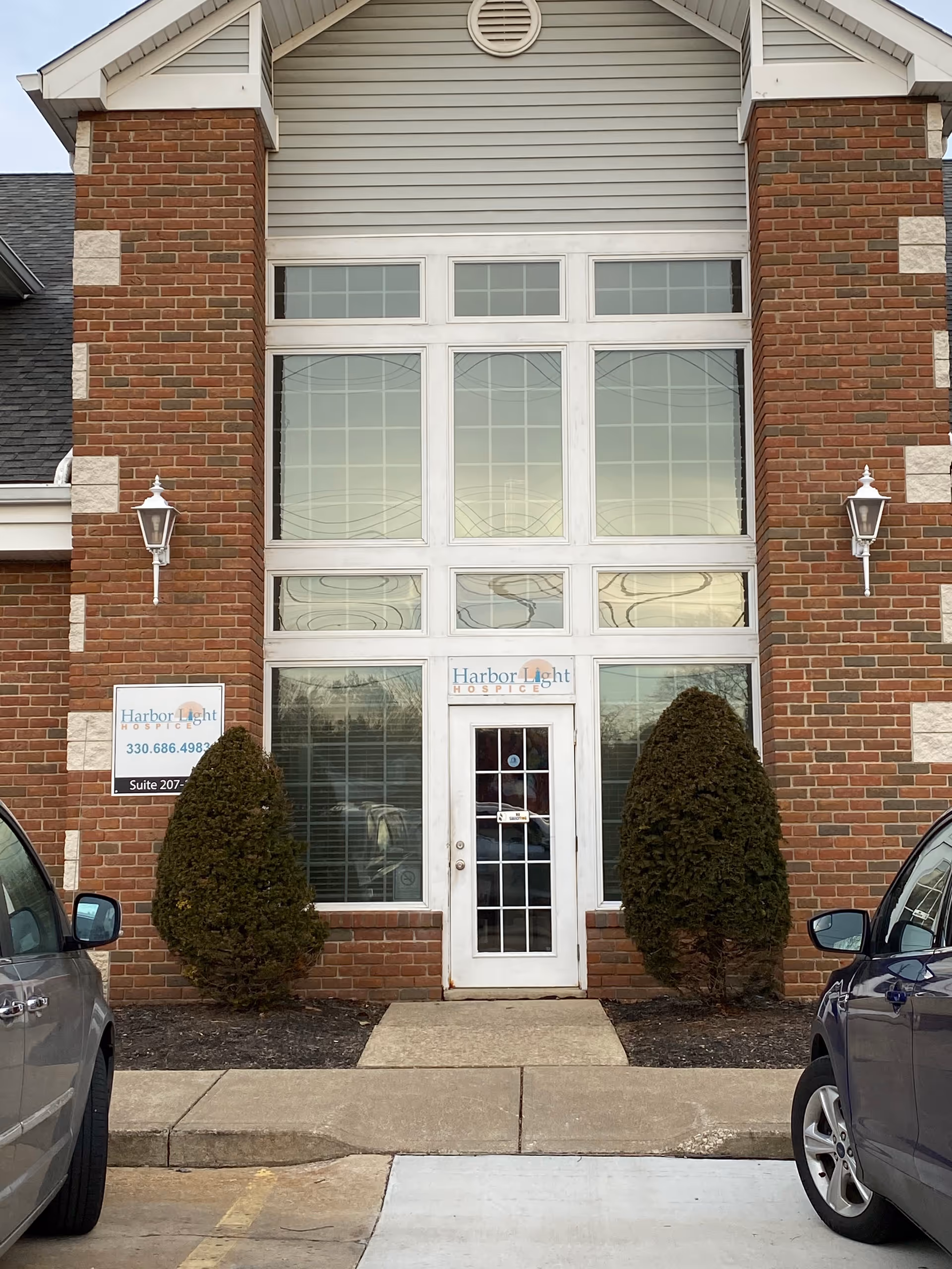 Brick building entrance with large glass doors, shrubs, and a small hospice sign.