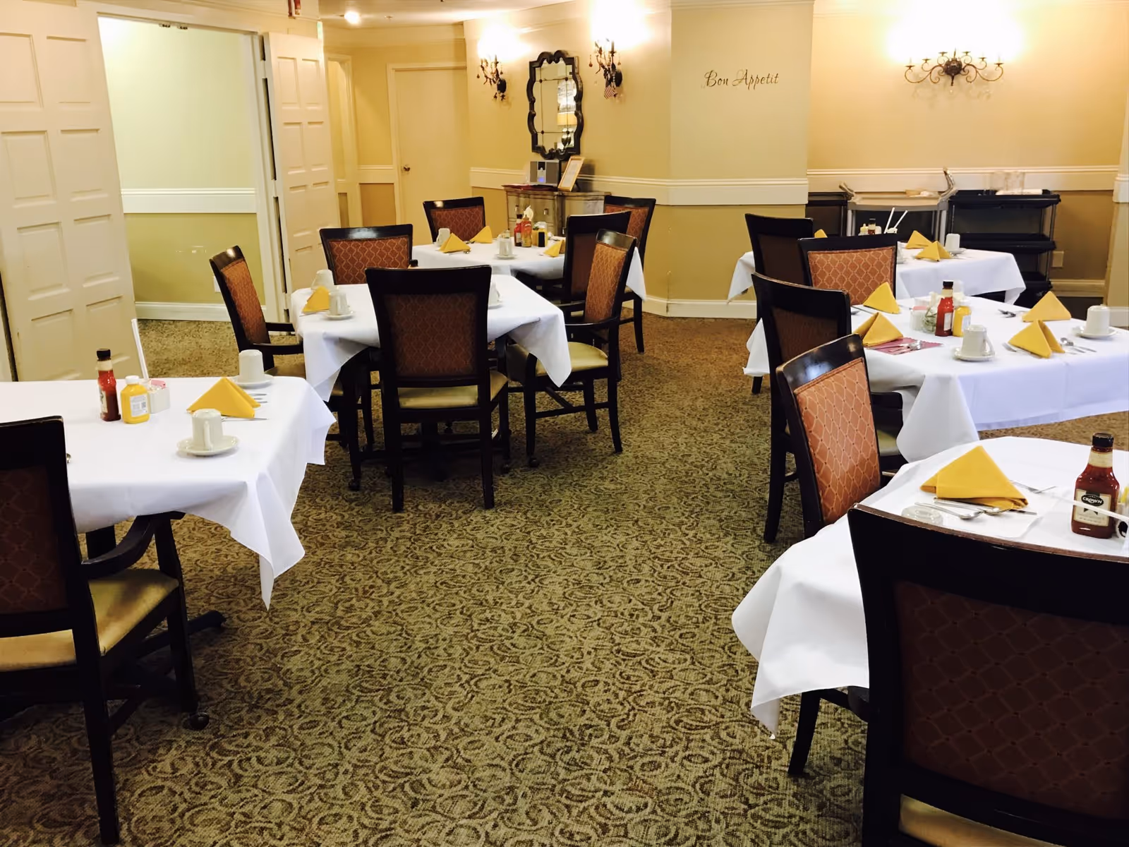 A dining room with several tables covered in white tablecloths, each set with yellow folded napkins, cups, and condiments. The room has patterned carpet, beige walls with decorative sconces, and a mirror on the far wall. The words 'Bon Appetit' are written on one wall.