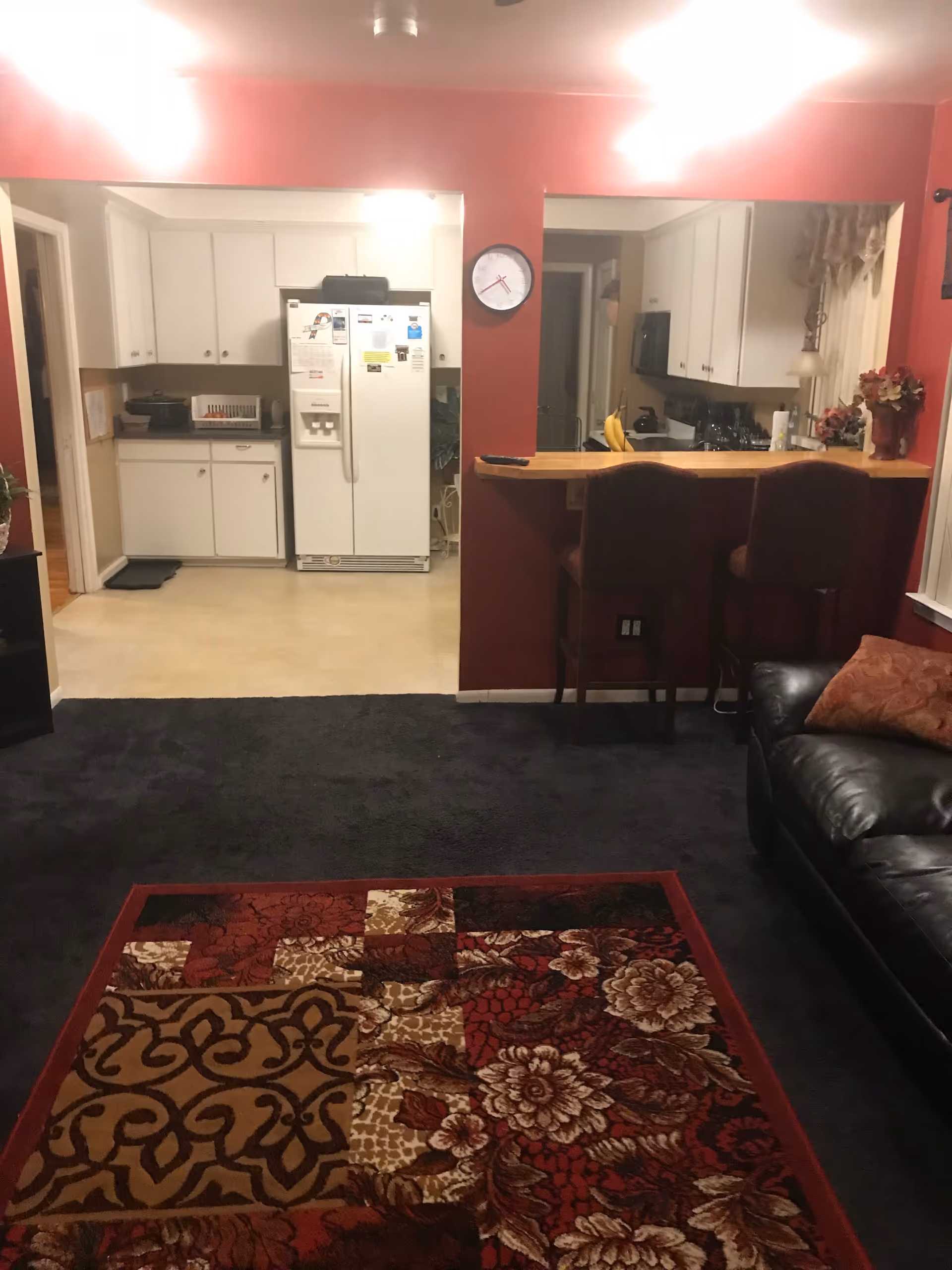 Cozy living room with dark carpet, leather sofa, patterned rug, and a kitchen bar.