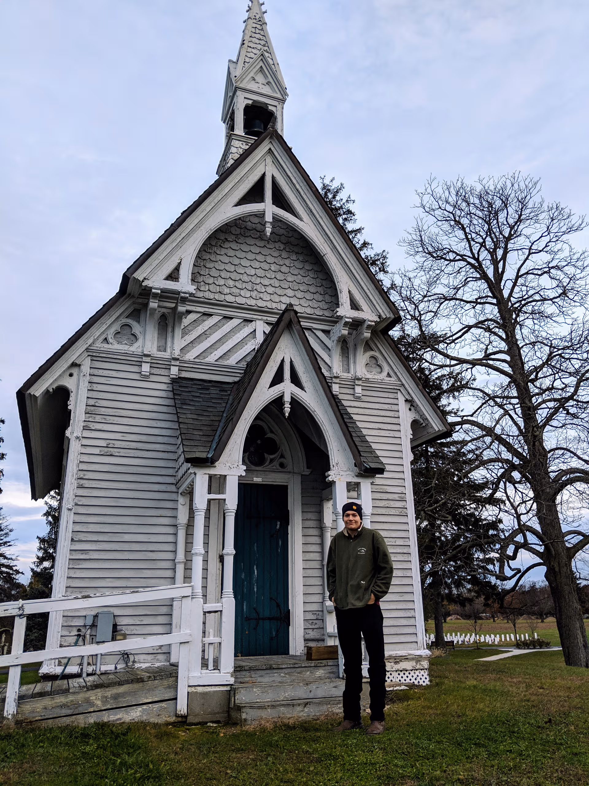 Small white wooden chapel with a man standing outside on grass.