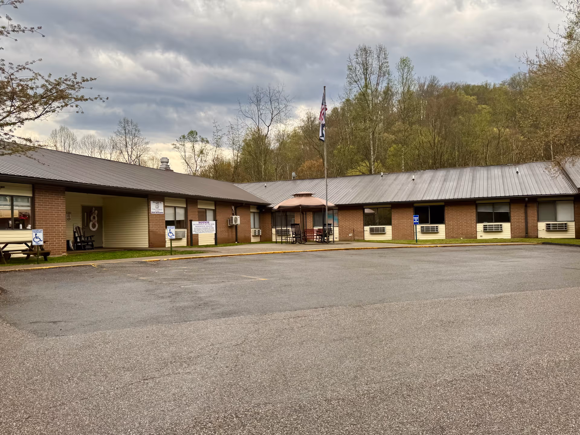 Single-story brick senior living facility exterior with parking lot, flagpole, and outdoor seating.