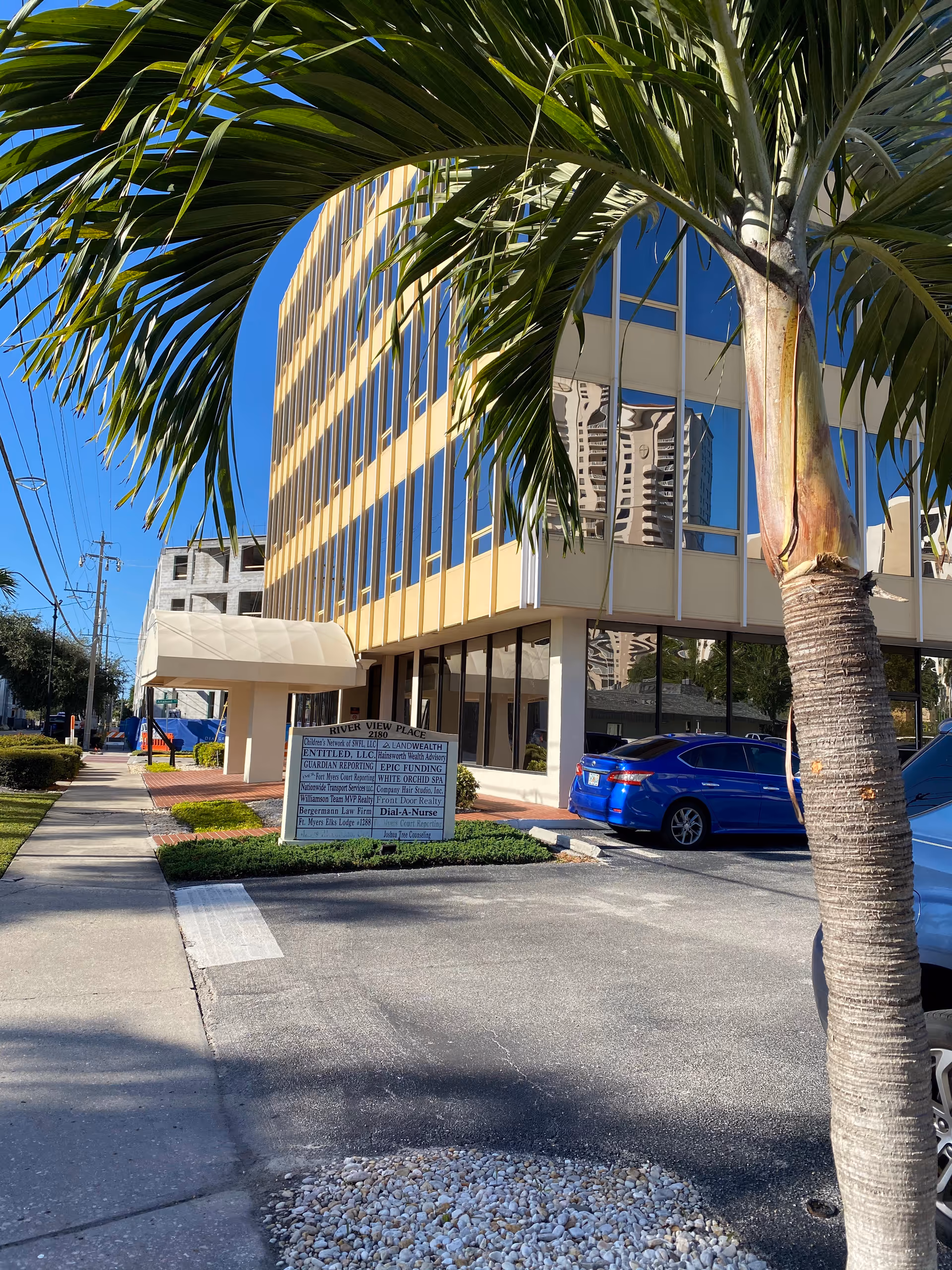 Exterior view of a modern building with glass windows, a palm tree, and a sidewalk sign.