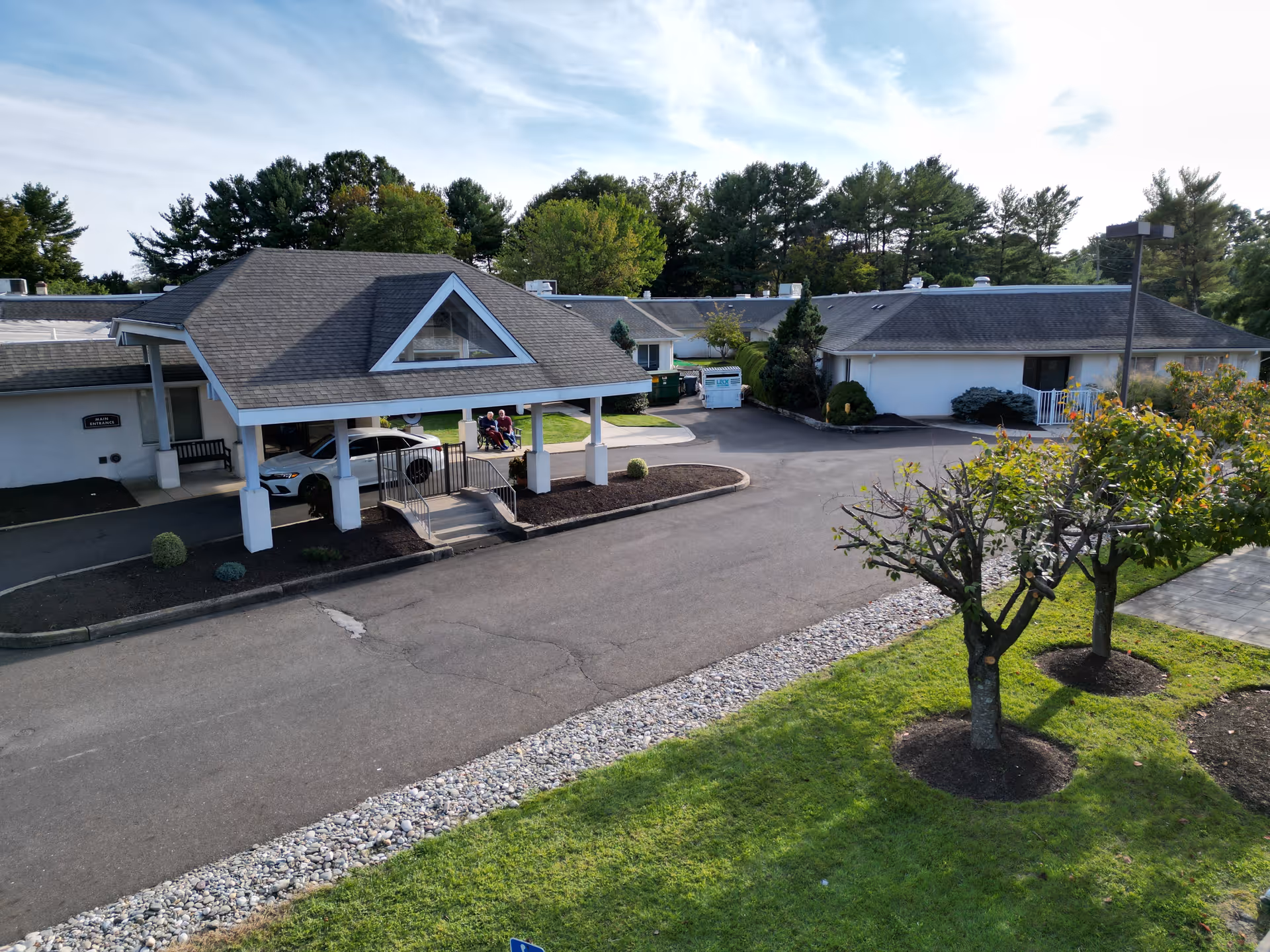 Exterior view of Chandler Hall Health Services building with a covered entrance, driveway, parked car, and a few people sitting outside. The area is surrounded by trees and greenery under a partly cloudy sky.