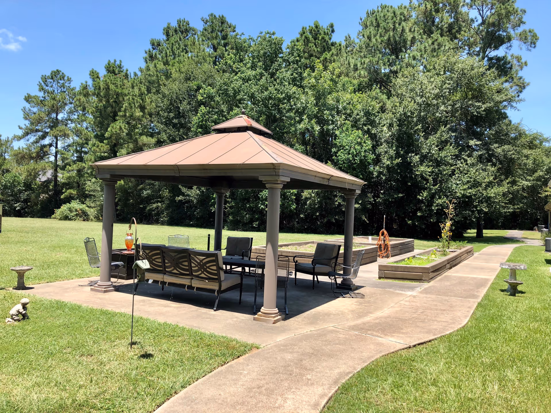 Outdoor gazebo with seating including cushioned benches and chairs on a concrete patio surrounded by green grass, raised garden beds, and trees under a clear blue sky.