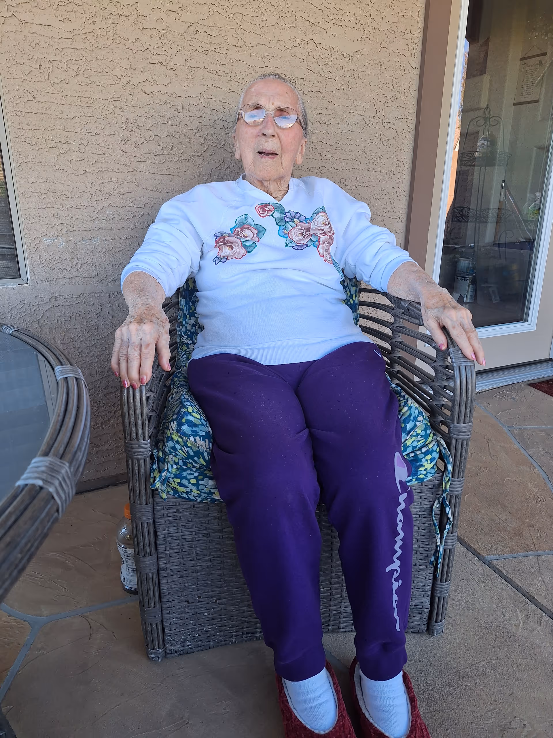 An elderly woman sits in a wicker chair on a patio outside a care home.