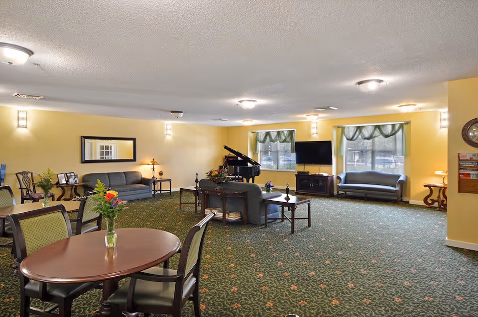 Senior living lounge with round tables, sofas, piano, TV, and large windows.
