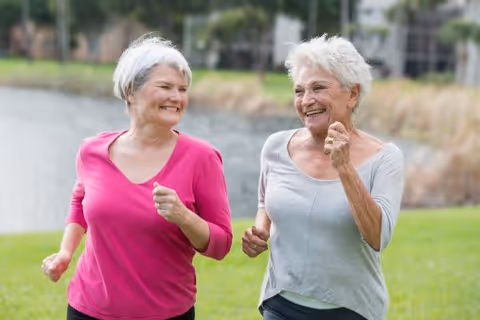 Two elderly women jogging outdoors on a grassy area near a body of water, both smiling and enjoying the activity.