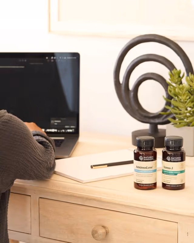 Wooden desk with a laptop, notebook, pencil, two supplement bottles, plant, and decorative sculpture.