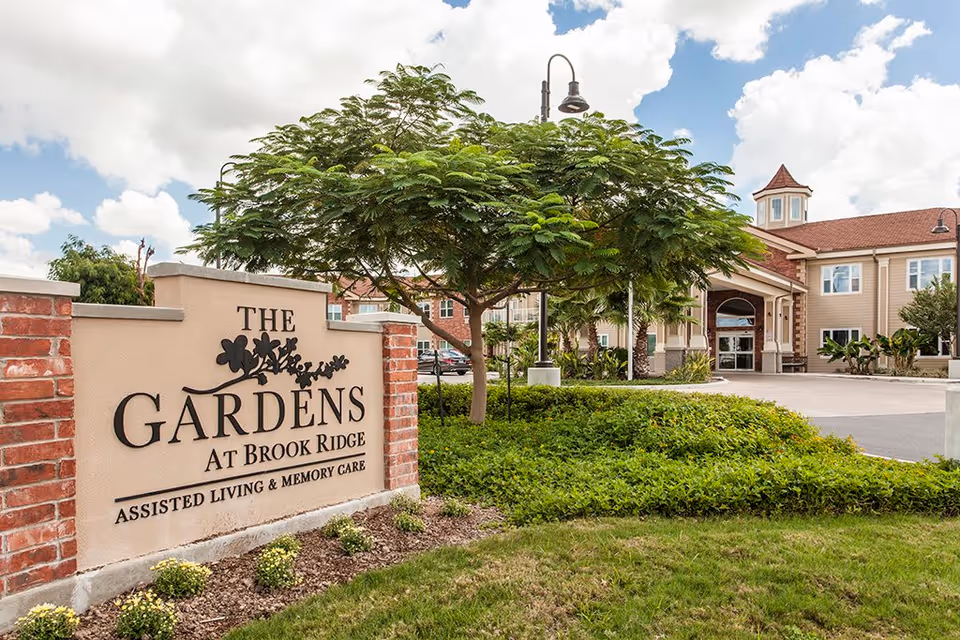 Entrance sign for The Gardens at Brook Ridge Assisted Living & Memory Care with a landscaped garden, a tree, and the facility building in the background under a partly cloudy sky.