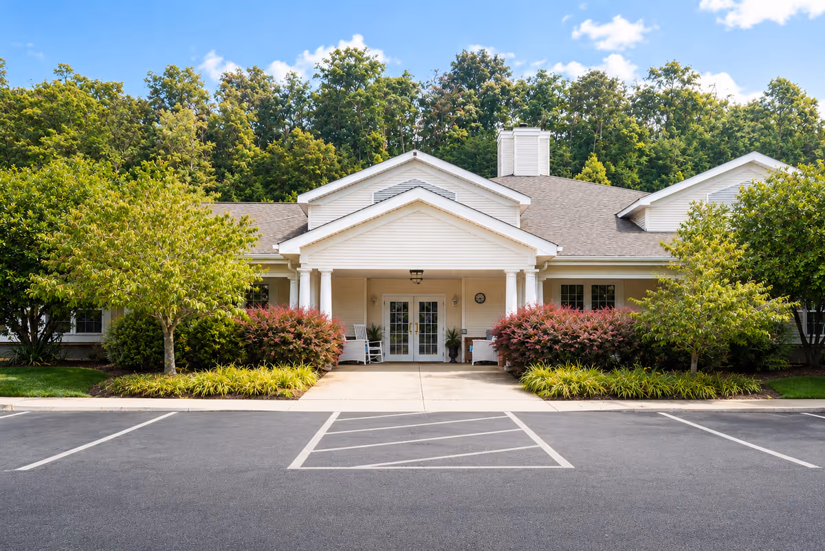 A beige senior living facility with a covered entrance, surrounded by trees and shrubs.