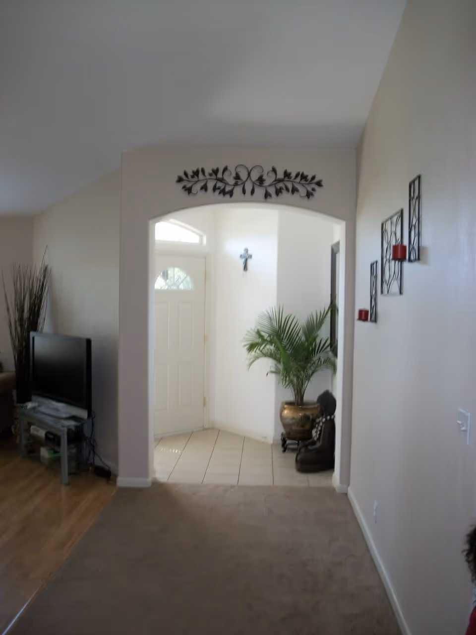 Interior view of a living space with a beige carpeted floor leading to a tiled entryway with a white front door. A decorative metal wall art is above the archway, and a potted plant and a small statue are placed near the door. On the left side, there is a TV on a stand with some items underneath, and on the right wall, there are three decorative candle holders with red candles.