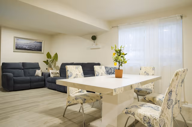 Bright senior living lounge with blue couches, a white dining table, and potted plants.