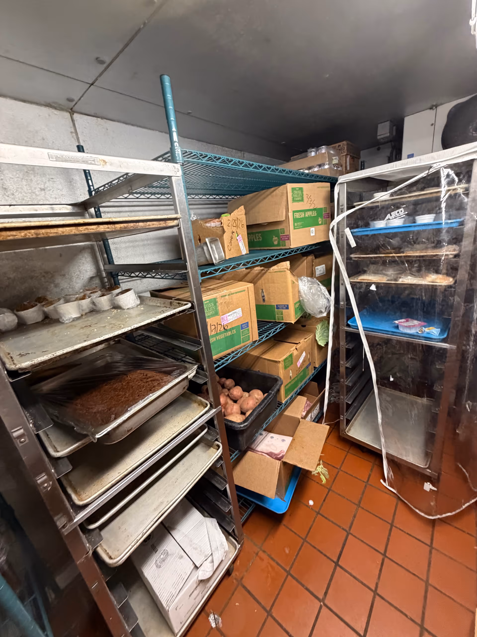 Industrial kitchen storage area with metal shelves, boxes, trays, and a glass-door cooler.