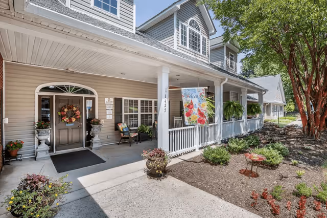 Front exterior view of a senior living facility named Navion of Shelby featuring a covered porch with seating, potted plants, a welcome flag, and well-maintained landscaping with trees and shrubs.