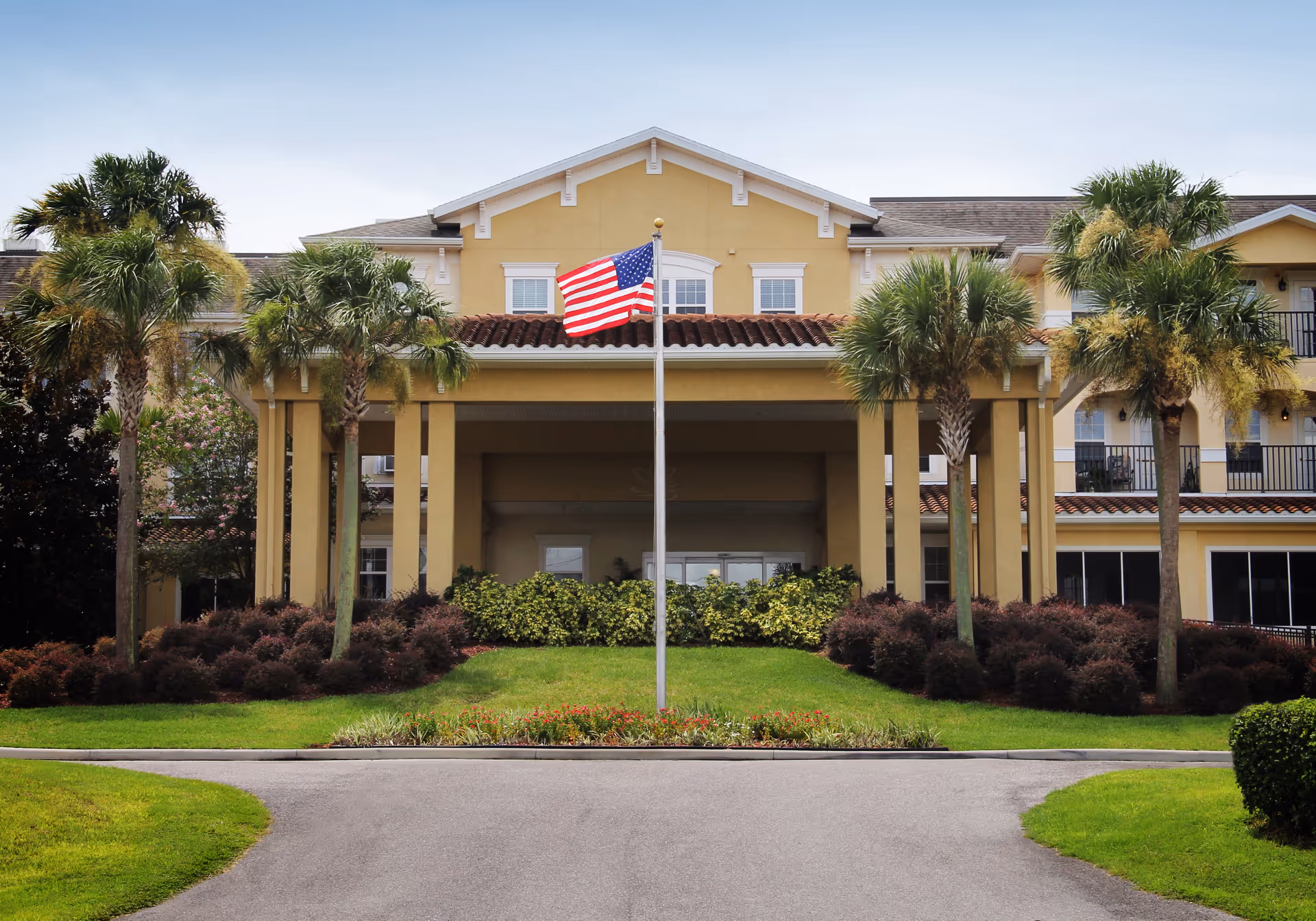 Front entrance of a two-story yellow building with an American flag, palm trees, and landscaped grounds.