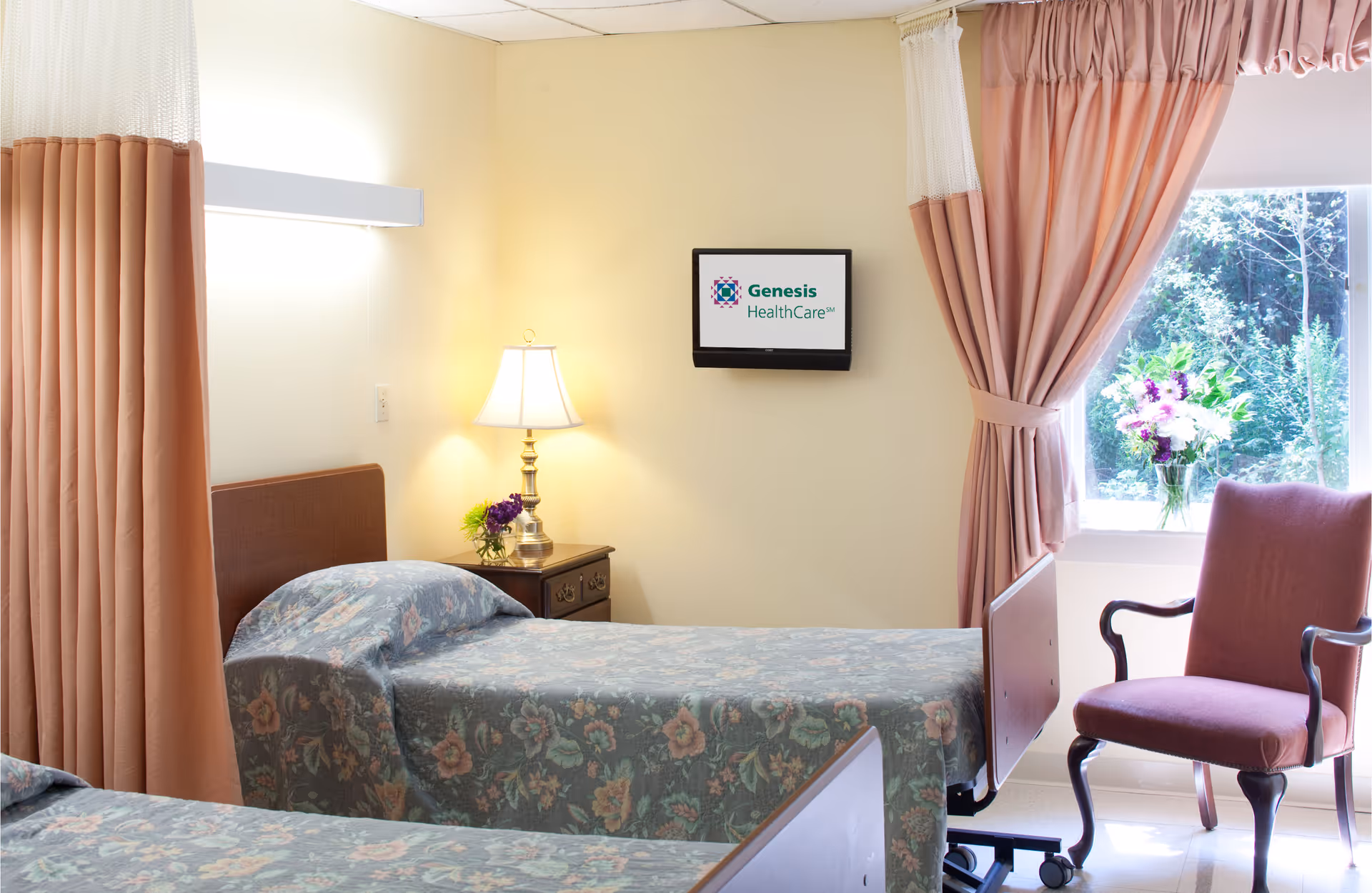 A bright rehab patient room with two floral-covered beds, a nightstand and lamp, a wall-mounted TV, and a window with curtains.