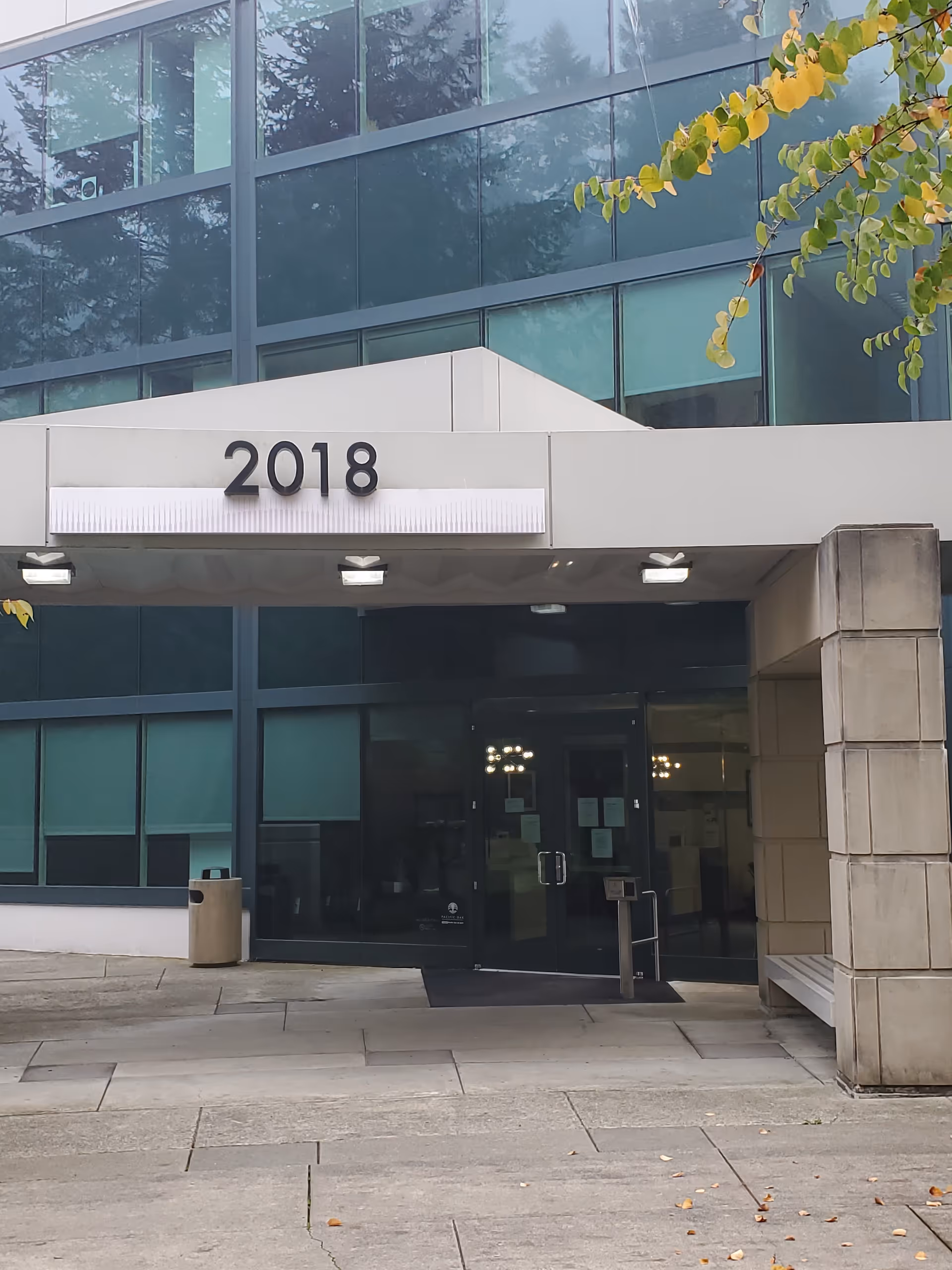 Modern glass-front building entrance with a 2018 sign, concrete columns, and glass doors.