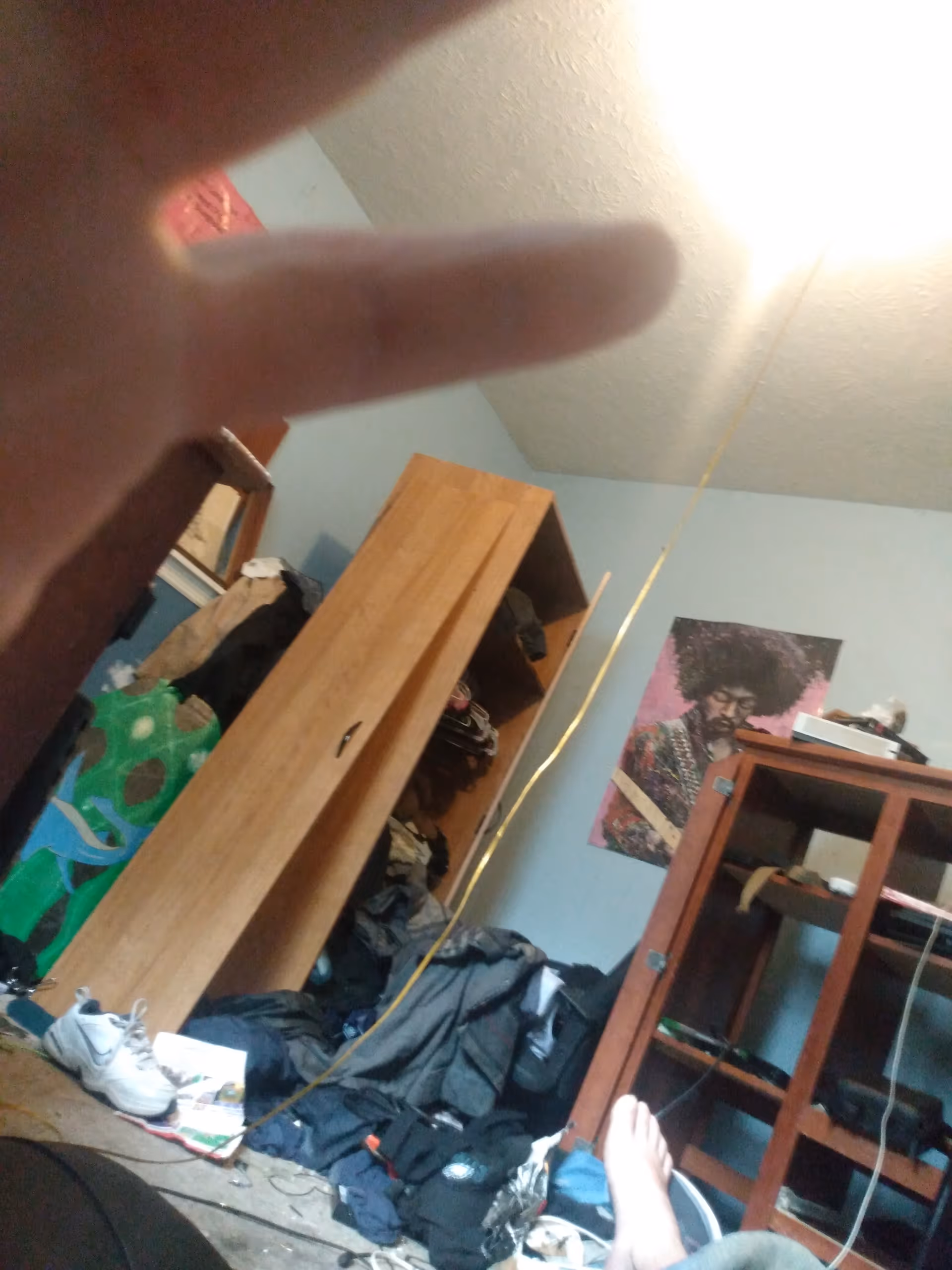 Cluttered bedroom with wardrobe, shelves, clothing on the floor, and a wall poster.