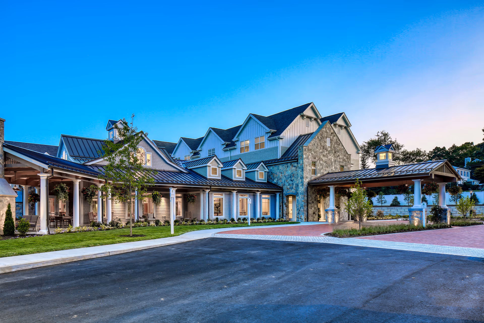 Exterior view of a large, modern senior living facility building with multiple peaked roofs, stone and siding walls, a covered entrance with columns, and well-maintained landscaping under a clear blue sky at dusk.
