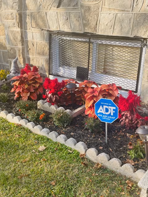 Exterior stone wall with window grate, colorful plants, and an ADT security sign