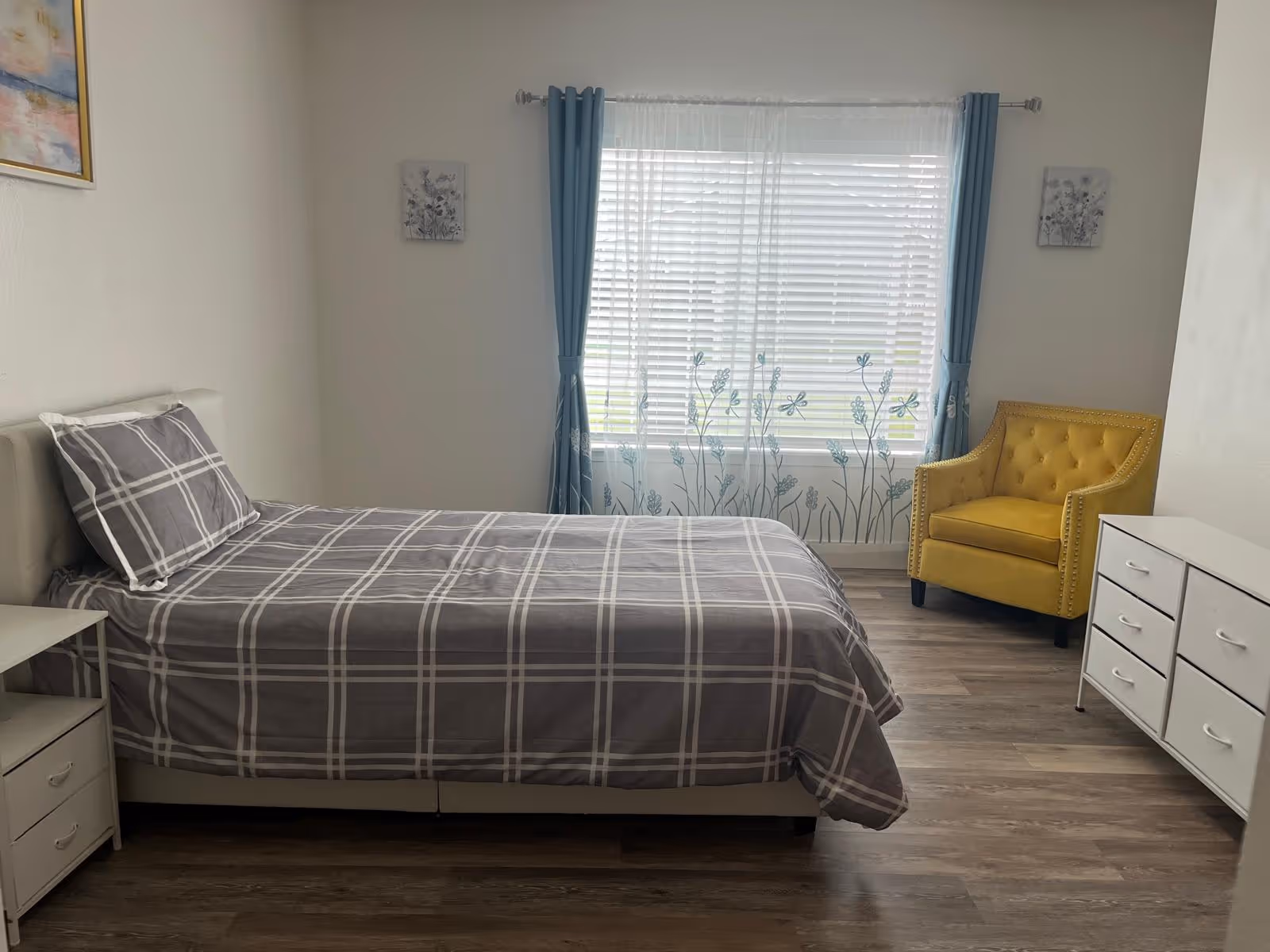 A furnished bedroom in a senior living facility with a bed, dresser, and yellow chair.
