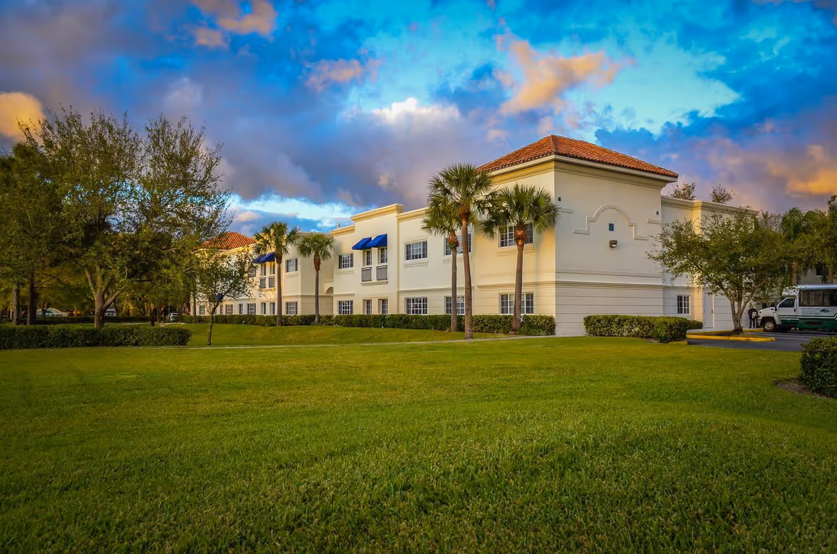 Cream-colored senior living facility exterior with manicured lawn, palm trees, and a blue sky.