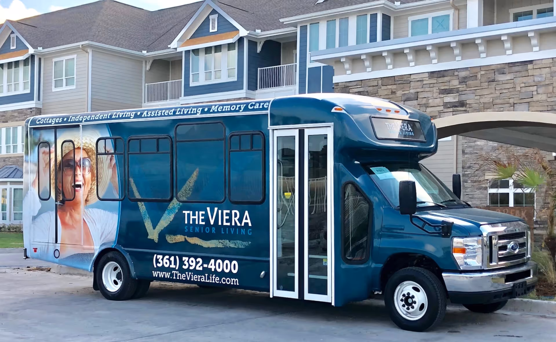 A blue shuttle bus parked in front of a senior living facility building. The bus has signage for The Viera Senior Living, including services like cottages, independent living, assisted living, and memory care. The bus features a large image of a smiling elderly woman wearing sunglasses and a sunhat.