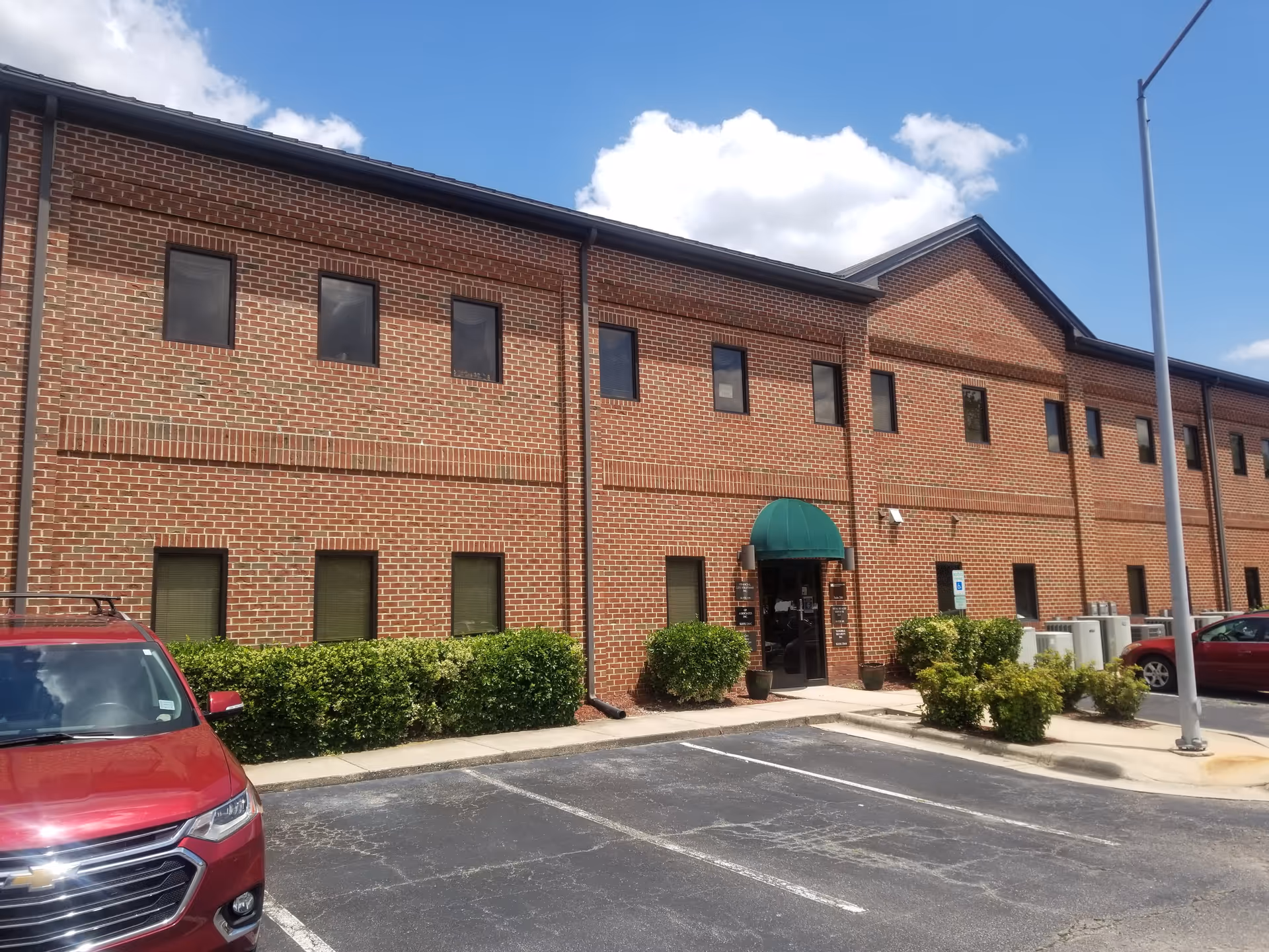 Brick senior living facility exterior with a green awning entrance, parking lot, and shrubs.