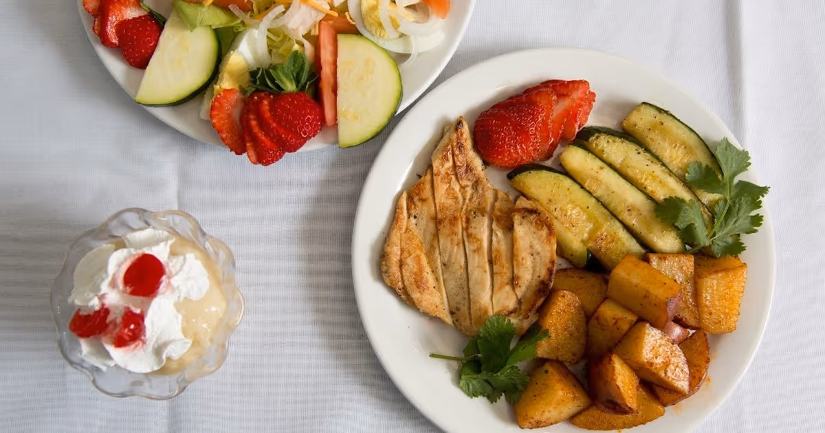 Plate of grilled chicken with roasted potatoes, sautéed zucchini and strawberries, accompanied by a salad and a dessert parfait on a white tablecloth.
