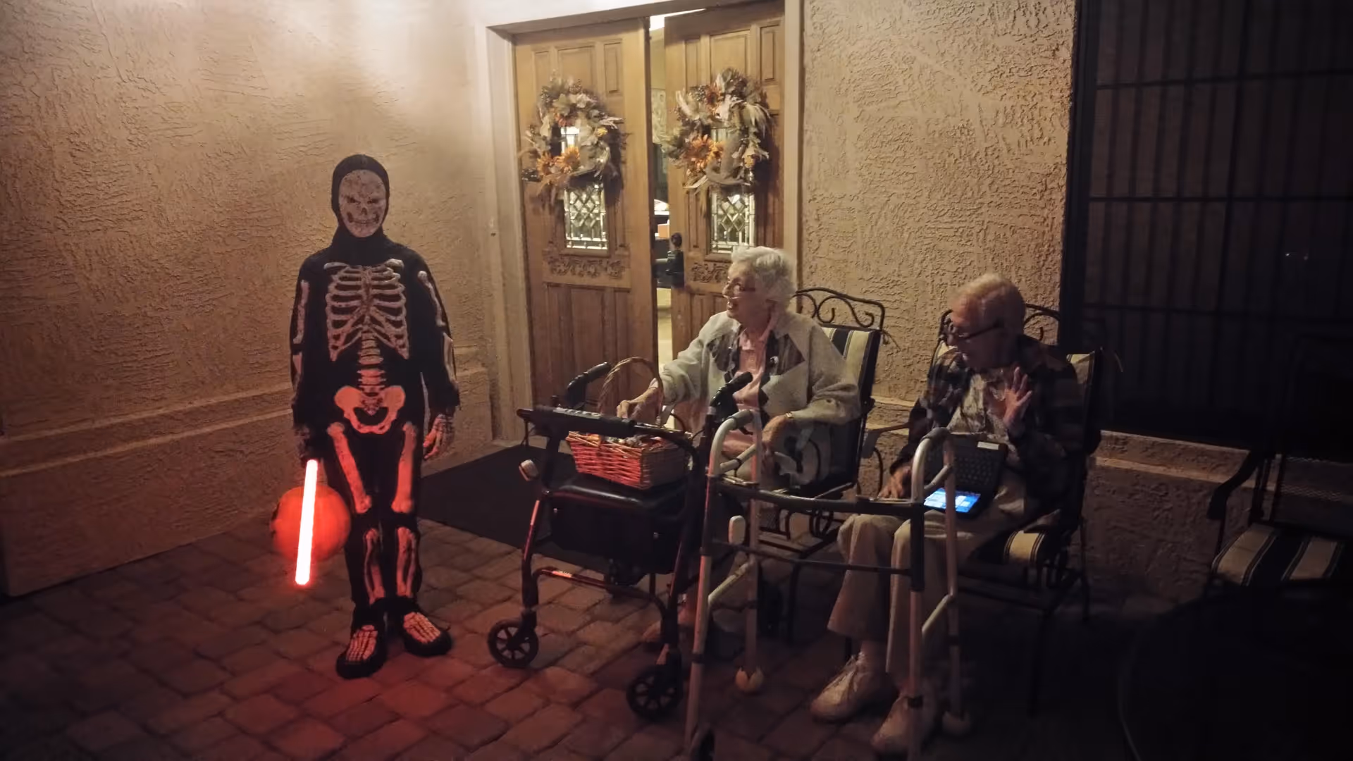 A person dressed in a glowing skeleton costume holding a light stick and a pumpkin bucket stands outside a building entrance decorated with autumn wreaths. Two elderly individuals sit nearby on chairs, each with a walker in front of them, engaging with the person in costume during nighttime.