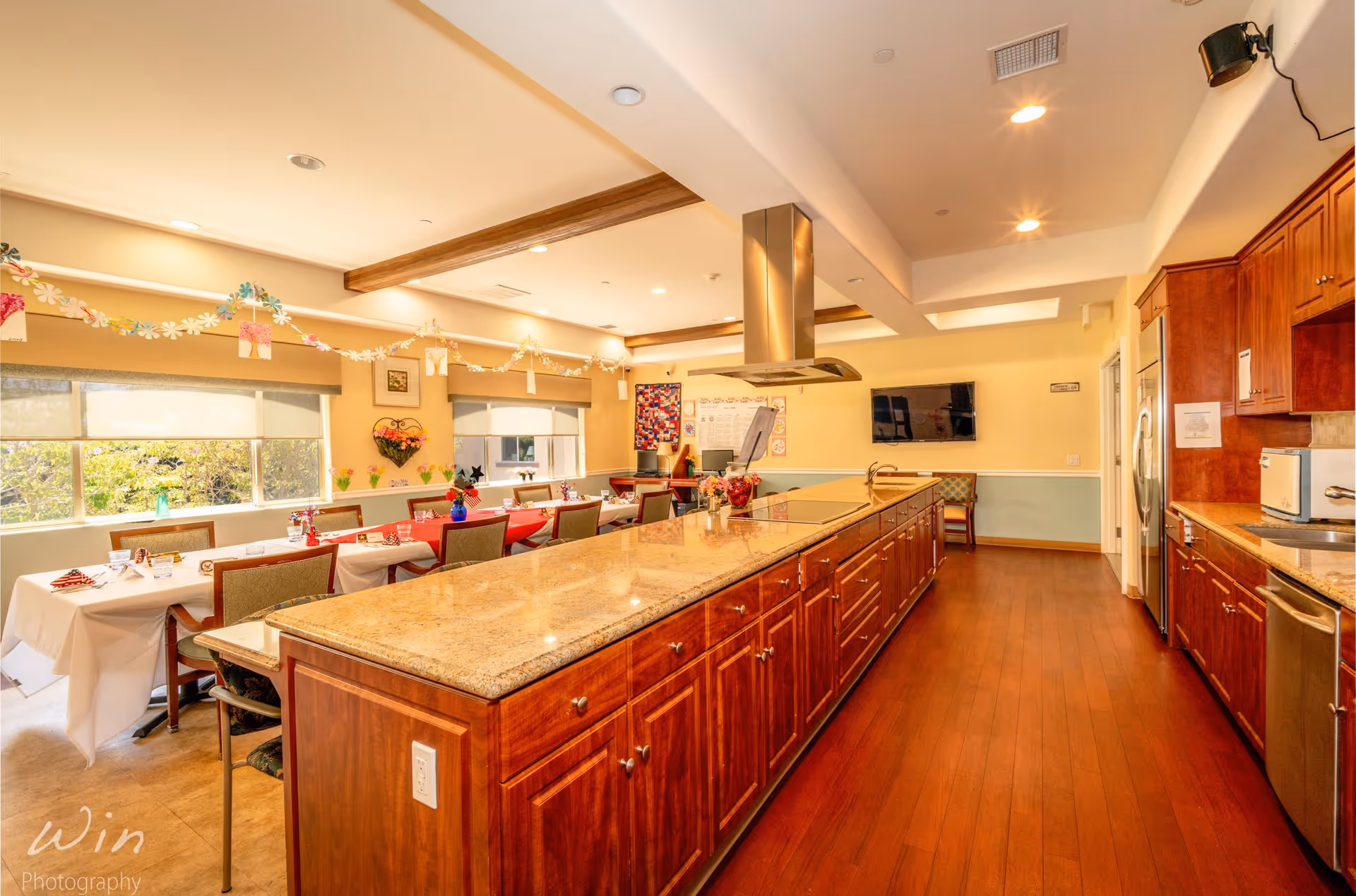 A bright and spacious kitchen and dining area in a senior living facility. The kitchen features a long granite countertop island with wooden cabinets, stainless steel appliances, and a range hood. The dining area has tables covered with white and red tablecloths, chairs, and decorative garlands hanging from the ceiling. Large windows let in natural light and provide a view of greenery outside.