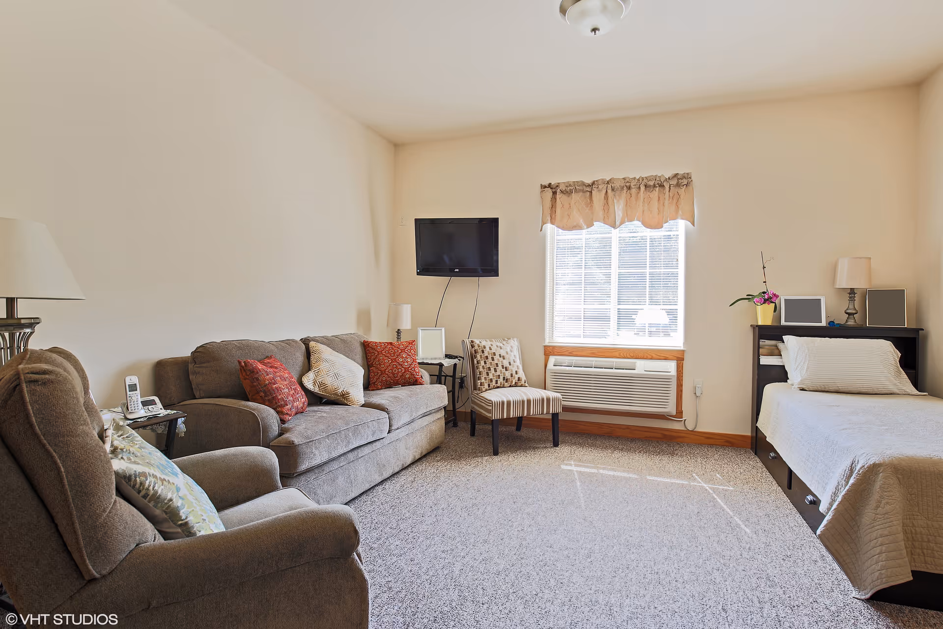 A cozy senior living suite with a sofa, chair, TV, window, and bed.