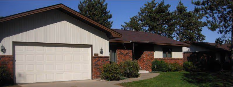Single-story brick and siding residential building with an attached garage, front lawn, and trees under a clear blue sky.