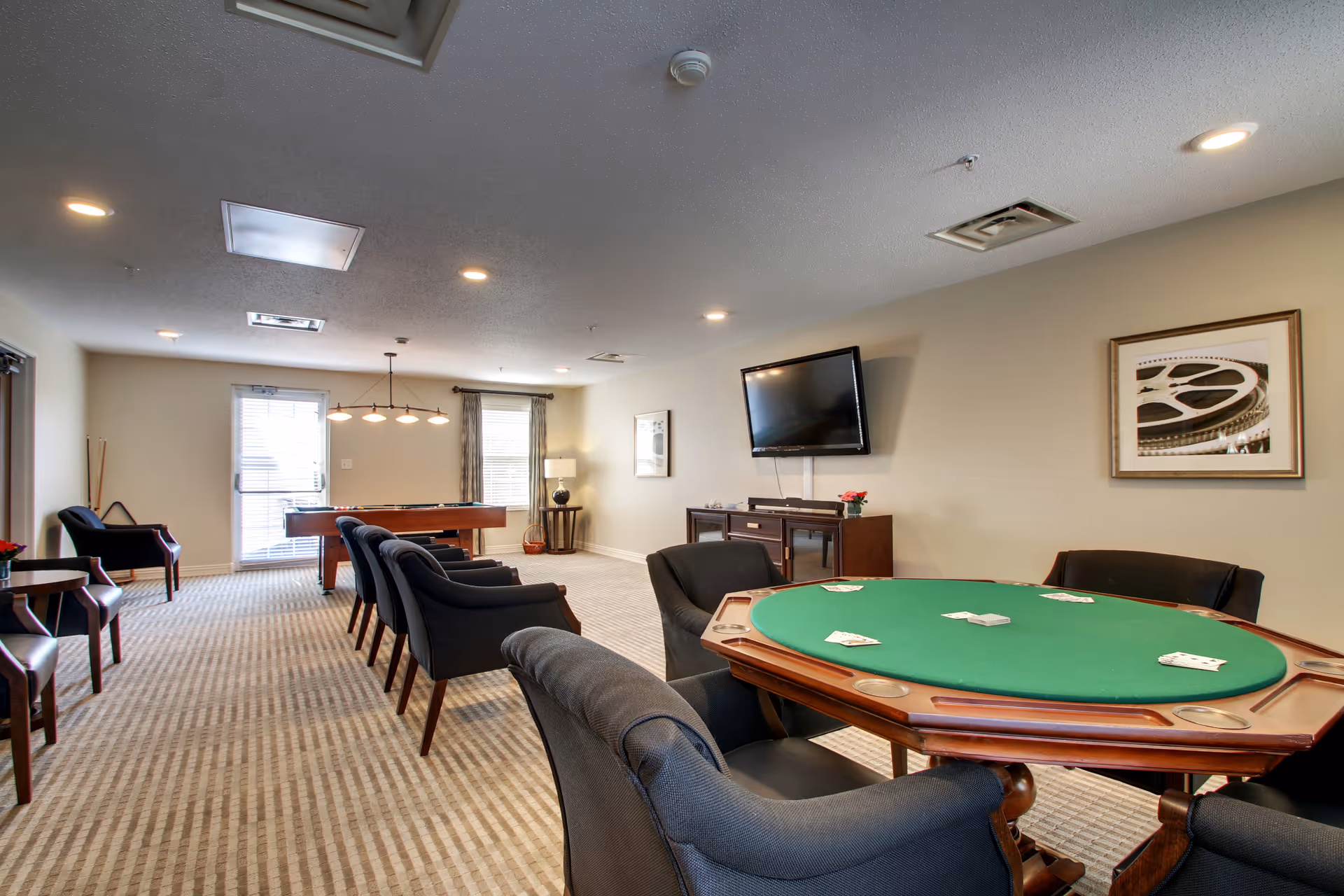 Sunlit senior living common area with round poker table, lounge chairs, and a wall-mounted TV.