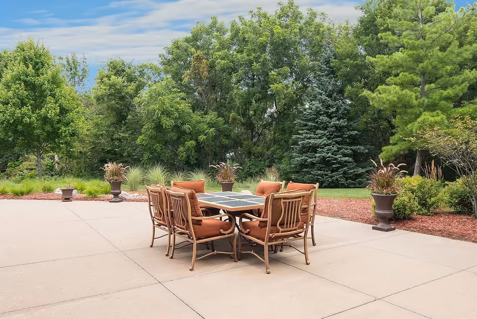 Outdoor patio area with a square table surrounded by eight cushioned chairs, set on a concrete surface. The background features lush green trees, shrubs, and decorative potted plants under a partly cloudy sky.