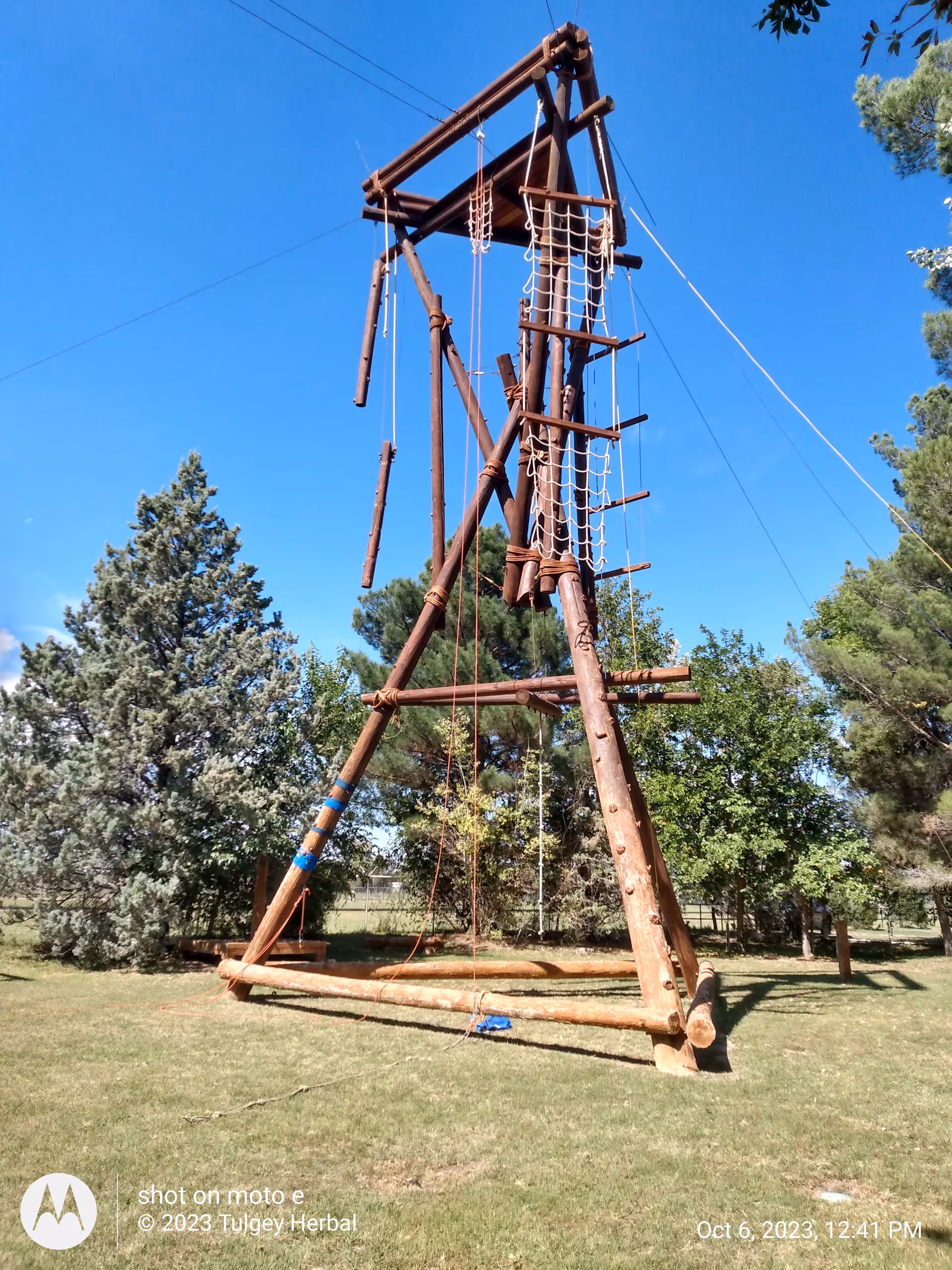 A tall wooden outdoor rope obstacle course stands on a grassy field with trees.