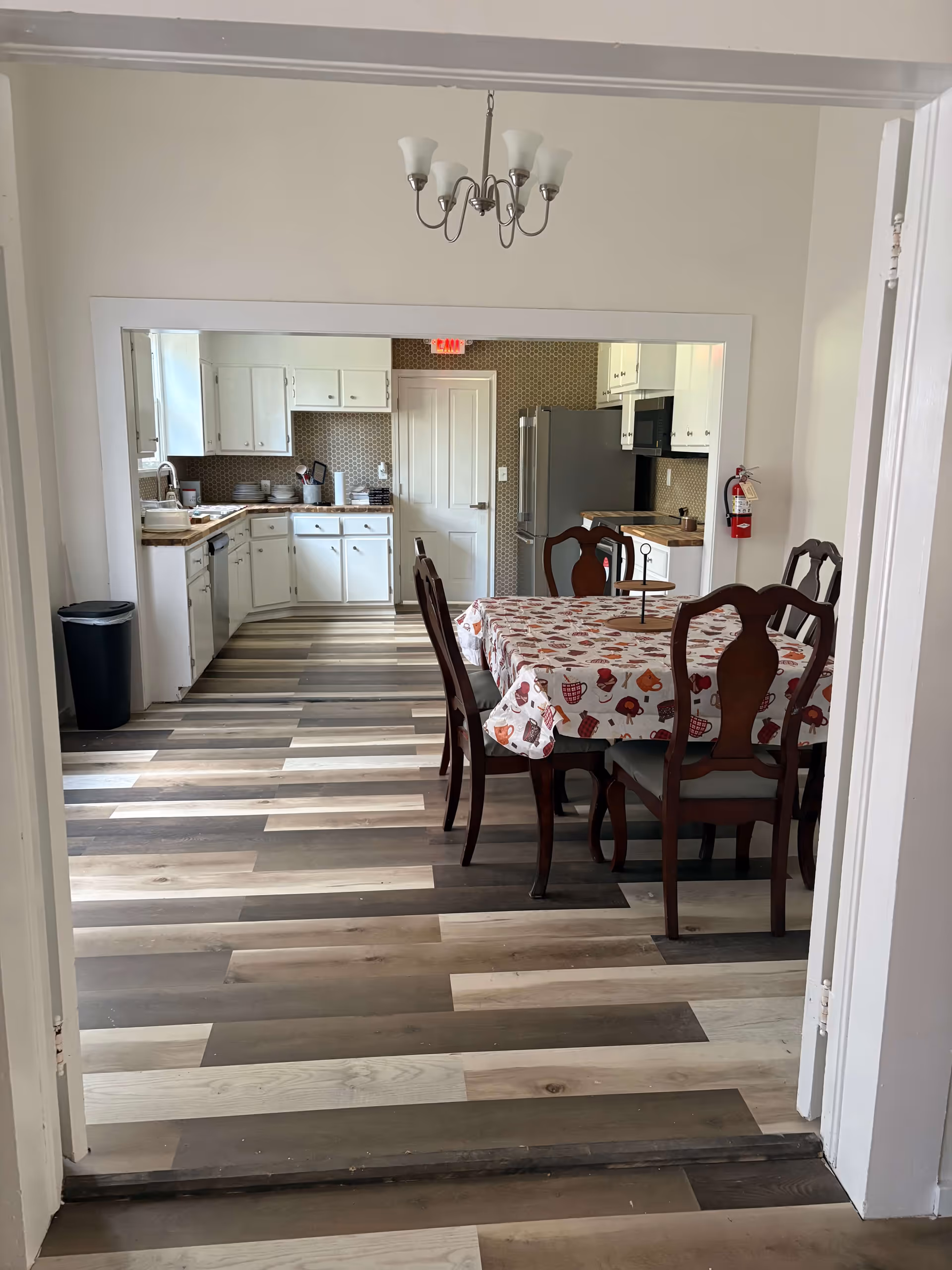 Dining room features a patterned tablecloth, wooden chairs, and a nearby kitchen.
