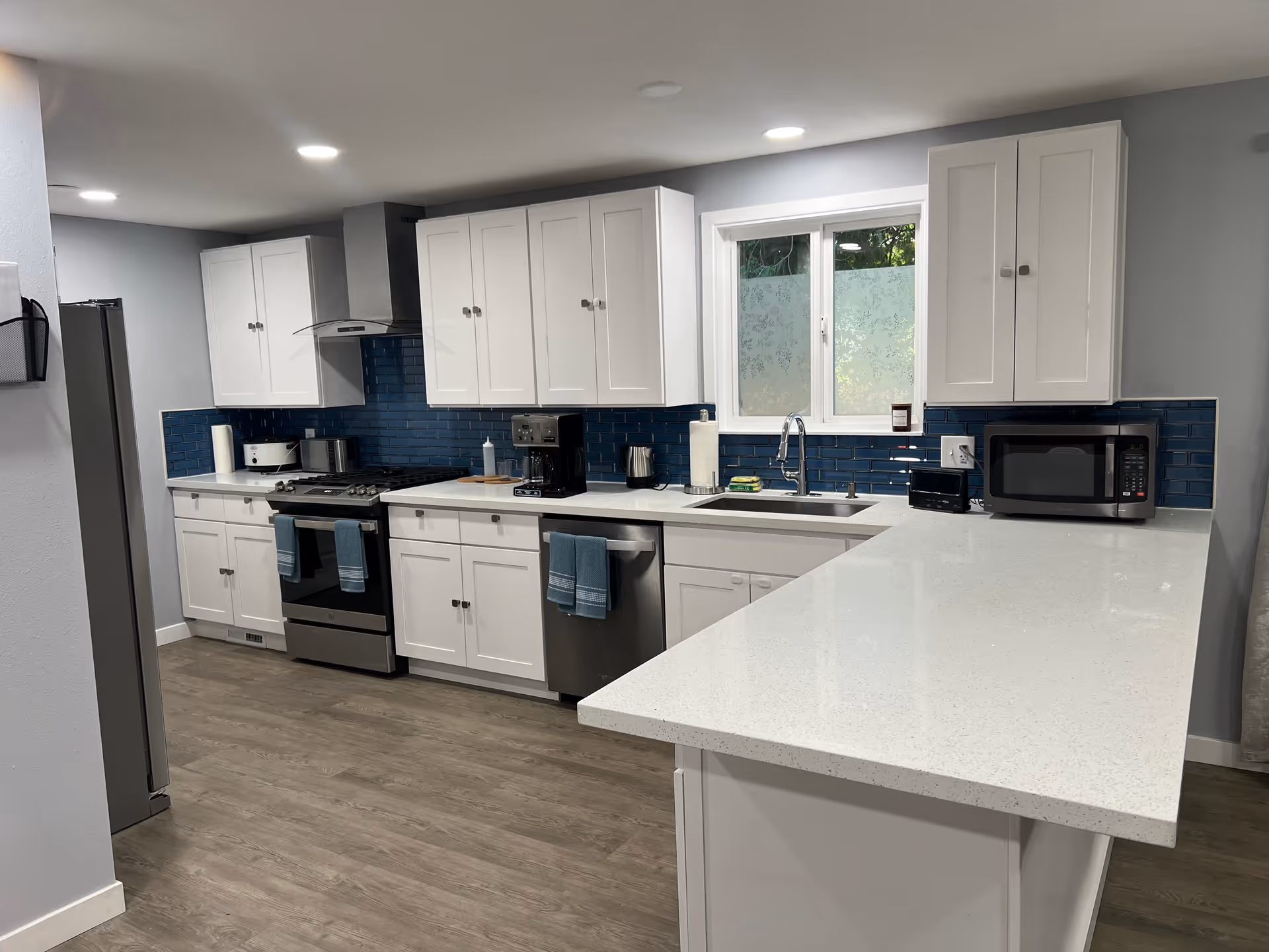Modern kitchen with white cabinets, a blue tile backsplash, stainless steel appliances including a refrigerator, stove, dishwasher, and microwave. There is a large white countertop island and a window above the sink.