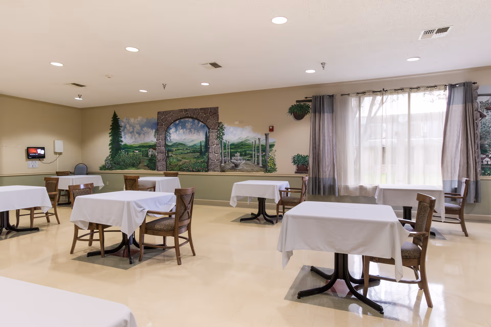 Dining room with multiple small tables draped in white cloths, wooden chairs, a mural on the far wall and a large curtained window.