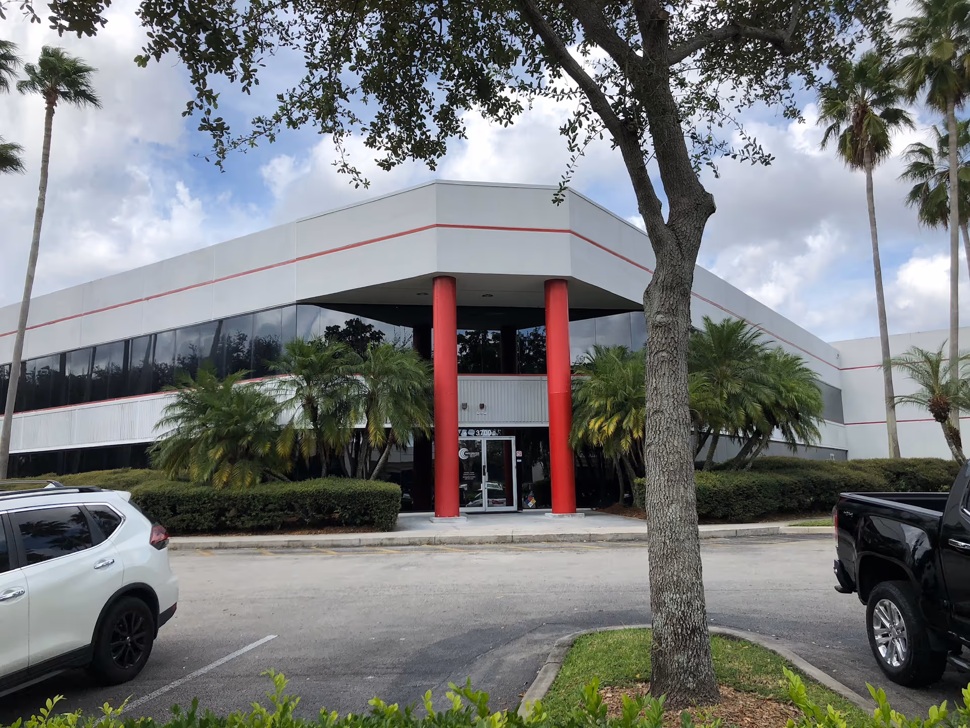 Modern single-story building with glass windows, red columns, palm trees, and parked cars.
