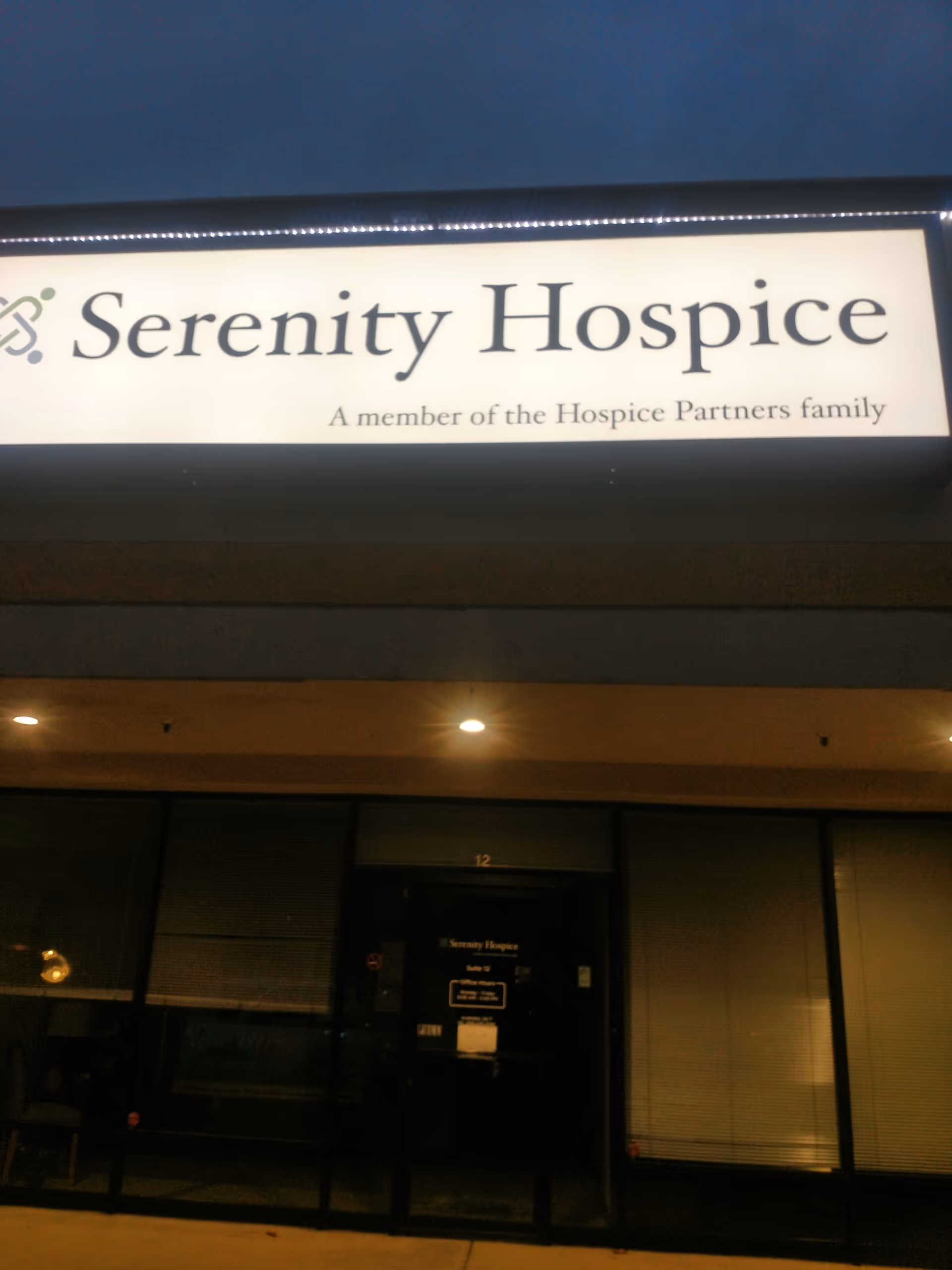 Nighttime storefront showing the Serenity Hospice sign above glass doors and windows.