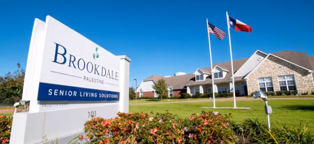 Exterior view of Brookdale Palestine senior living facility with sign, flags, and manicured lawn.