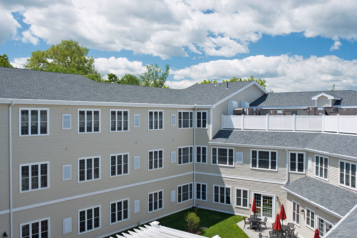 Exterior courtyard view of a multi-story beige senior living building with many windows and a small patio area with tables and red umbrellas under a blue sky.