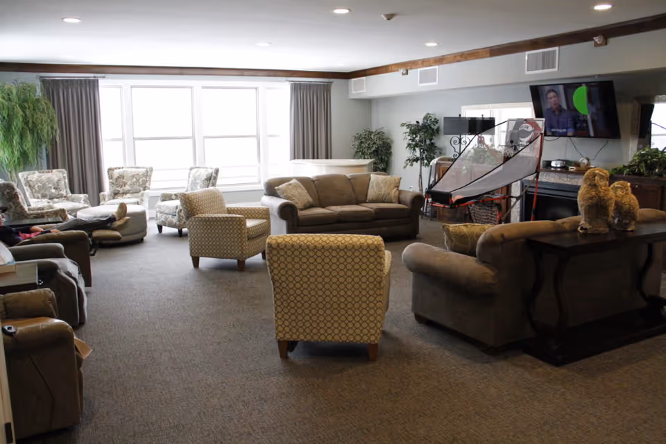 A spacious senior living lounge with sofas, chairs, plants, and a mounted TV.