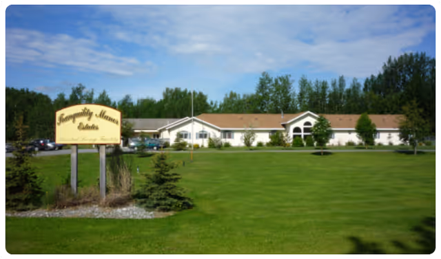 Single-story senior living building seen across a large green lawn with a freestanding entrance sign in front and trees behind it.