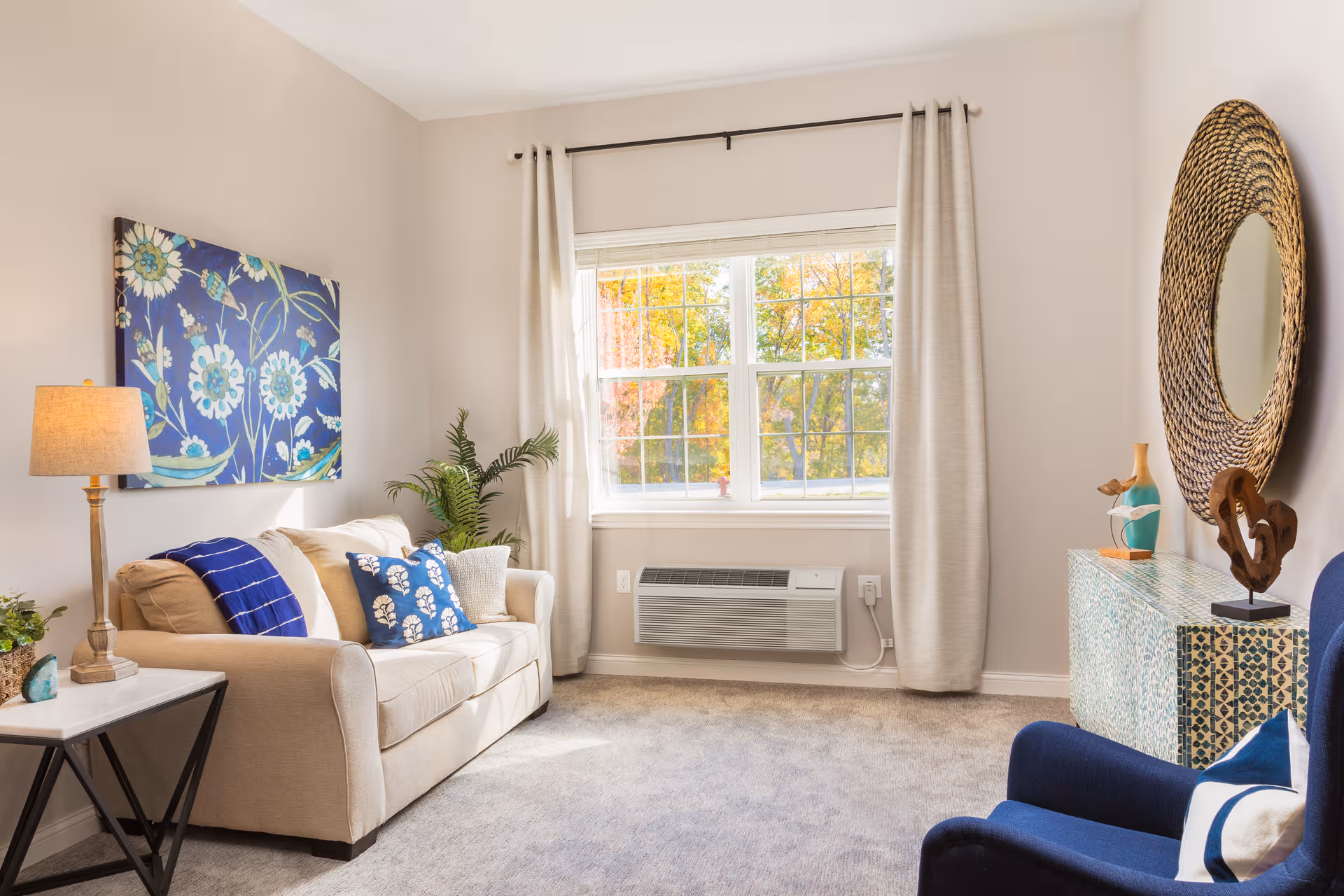 Bright living room with beige sofa, blue accents, plants, and a round mirror.